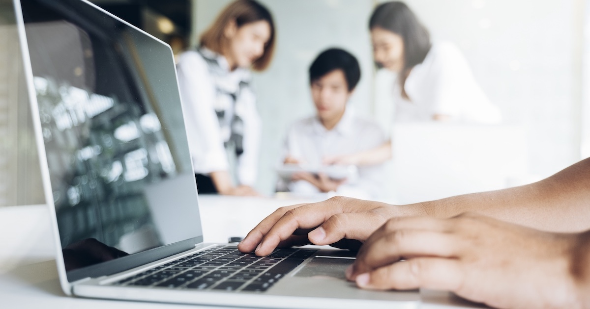 パソコンに手を置くビジネスパーソンと背景にミーティングをする人達