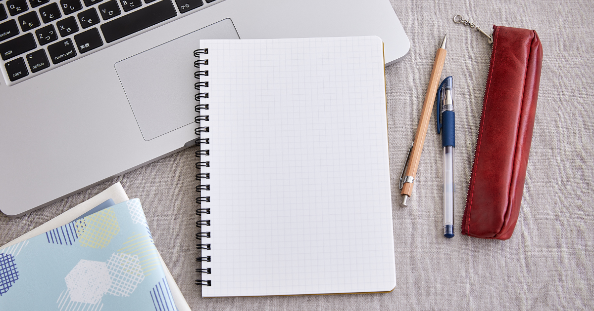 方眼ノート、赤いペンケースのそばにシャープペンシルとボールペン、手帳、ノートパソコンが置いてある