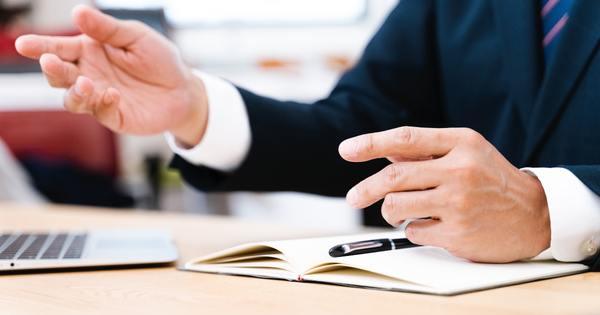 手元にノートとノートパソコンを置き、プレゼンをしている、スーツ姿の男性の手