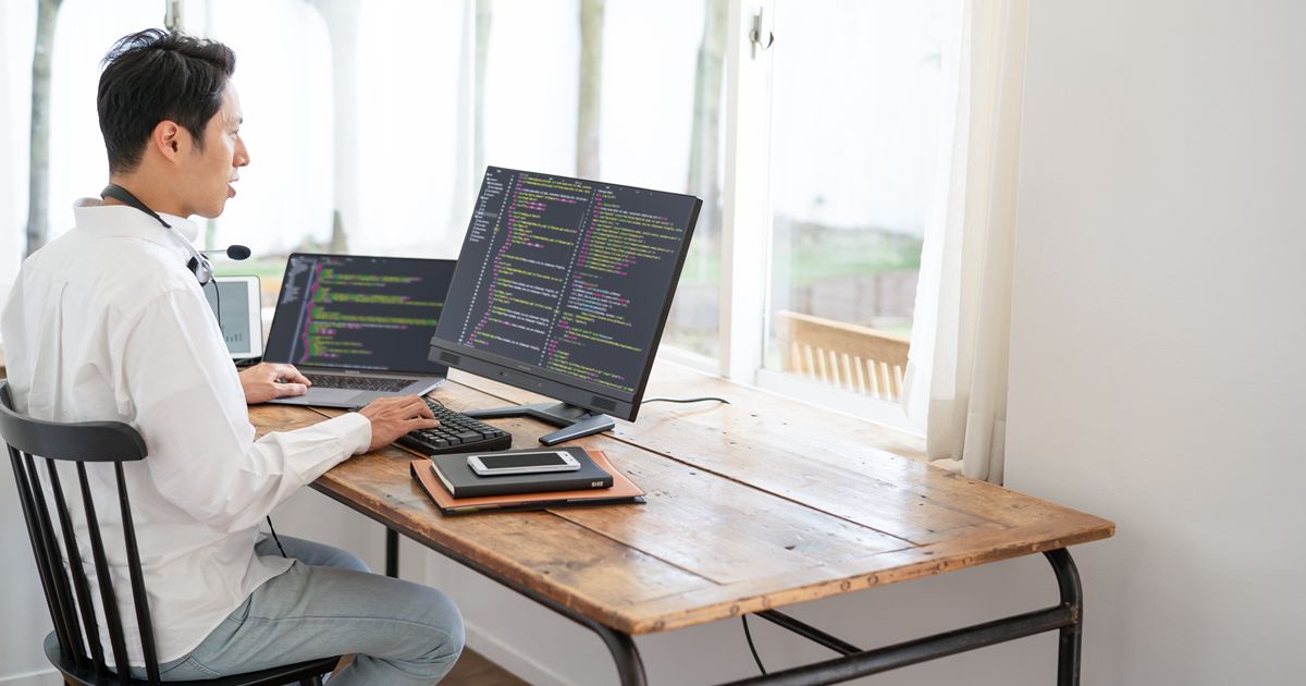 自宅のデスクでデュアルモニターを使ってプログラミングをする男性