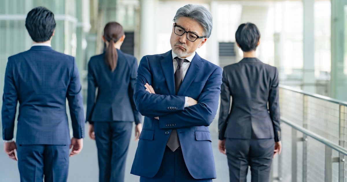 若いビジネスパーソン達を背に、腕を組み考える白髪のビジネスパーソン