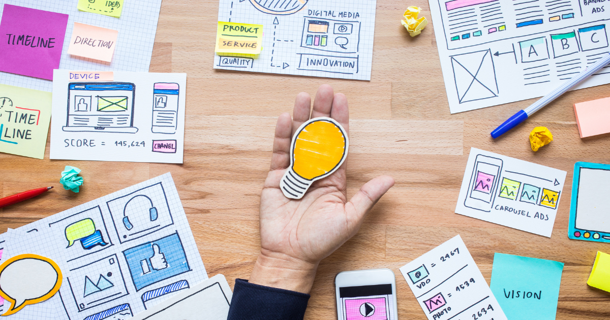 Webデザインのラフや付箋が並ぶ机の上で、ひらめきを象徴する電球アイコンを手に持っている様子