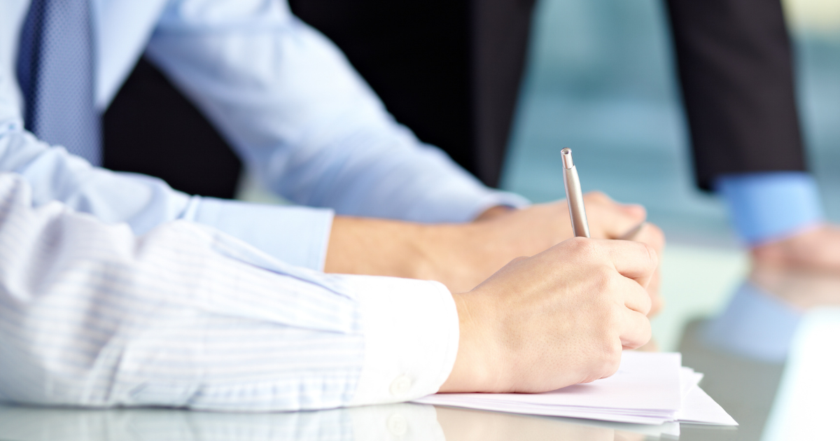 ノートを書くワイシャツ姿の男性の手元、背景にスーツ姿の男性の手元