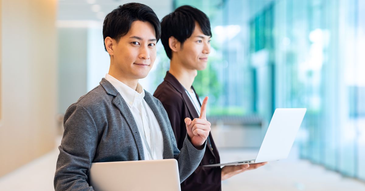 ノートパソコンを持ち笑顔で立つ男性と指を立てる同僚