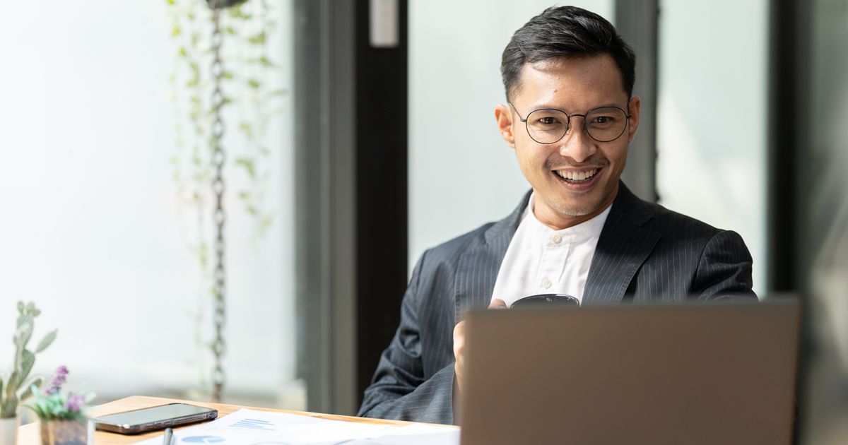 オフィスでコーヒーを飲みながら笑顔でノートパソコンを見るスーツ姿の男性