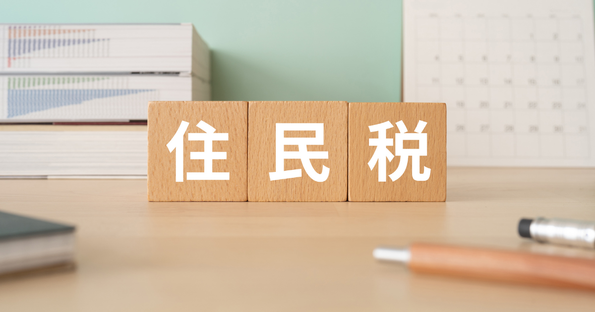 「住民税」と書かれた木製ブロックが手帳とペンのそばに置かれており、背面に本とカレンダーが置いてある