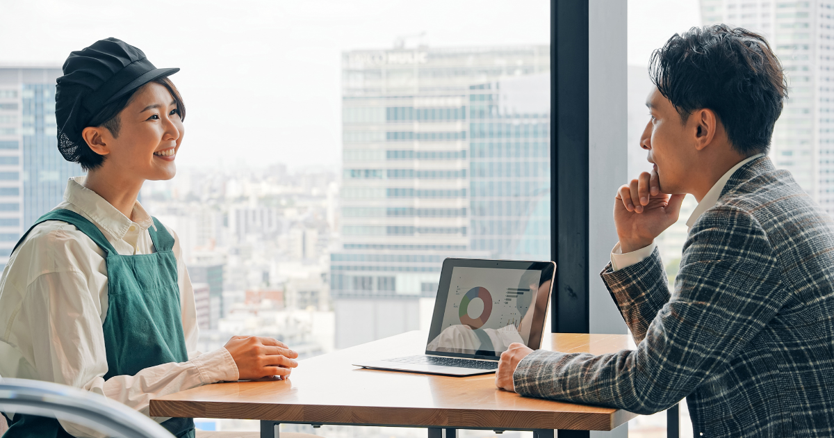 窓際のテーブルで、男女がノートPCを挟んで向かい合って融資の相談をしている