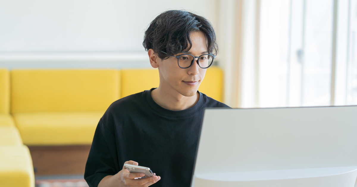 ノートパソコンの前に座り、スマートフォンを見ながら作業をする男性の様子