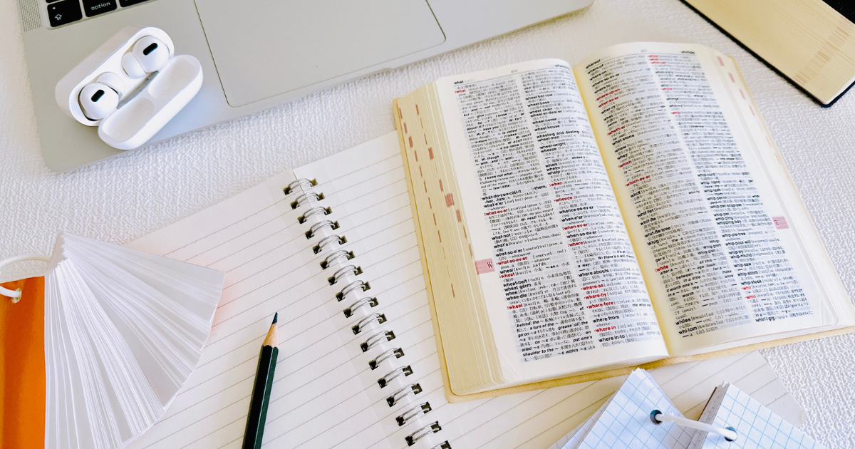 開いた本とリングノートがあり、ワイヤレスイヤホンや鉛筆、ノートPCの一部、本が写っている