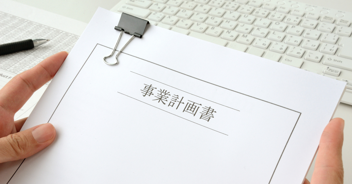 クリップで留めた書類「事業計画書」を両手で持っている