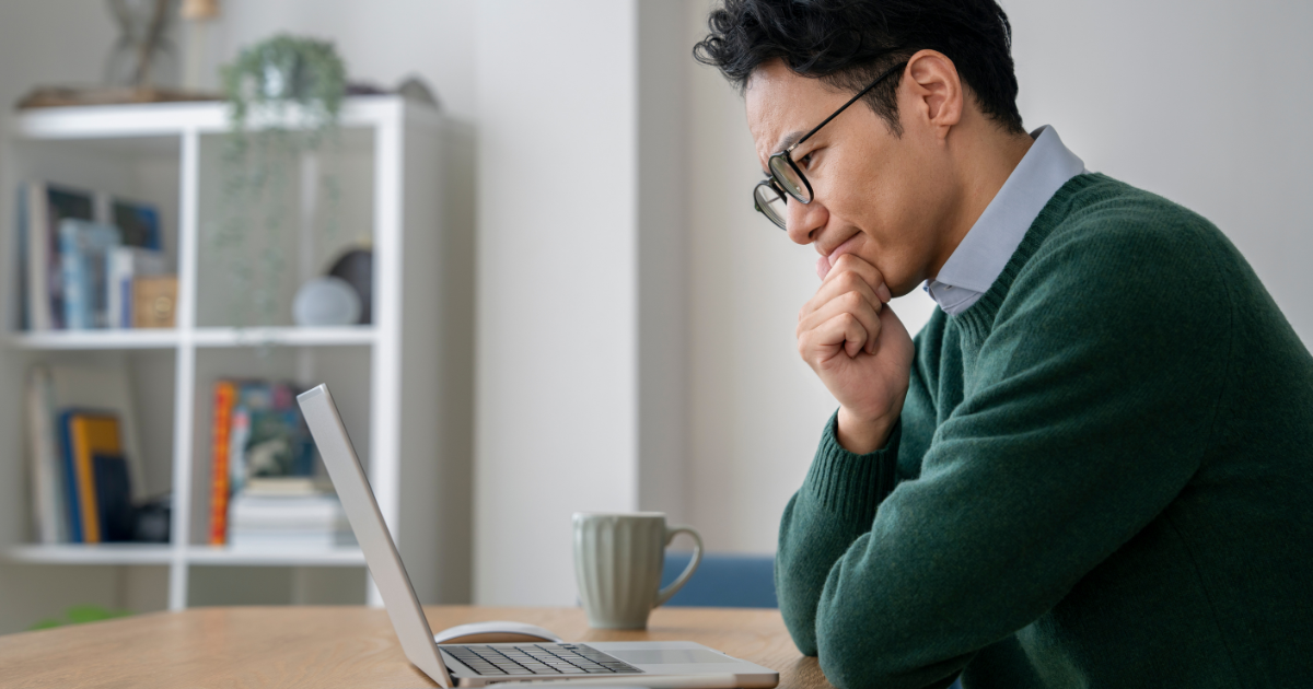 背後に棚がある部屋で、ノートPCの前で考え込んでいる眼鏡の男性