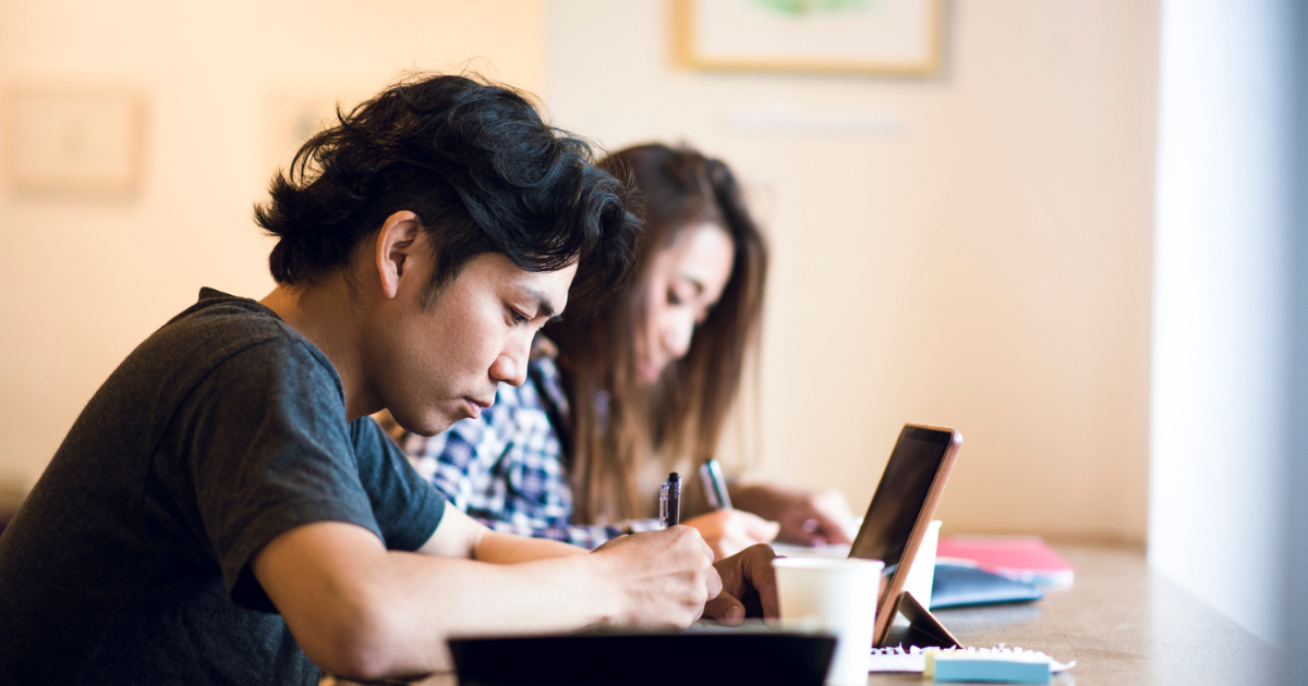 机に向かって並び、ノートパソコンと書類を見て仕事をしている様子の二人