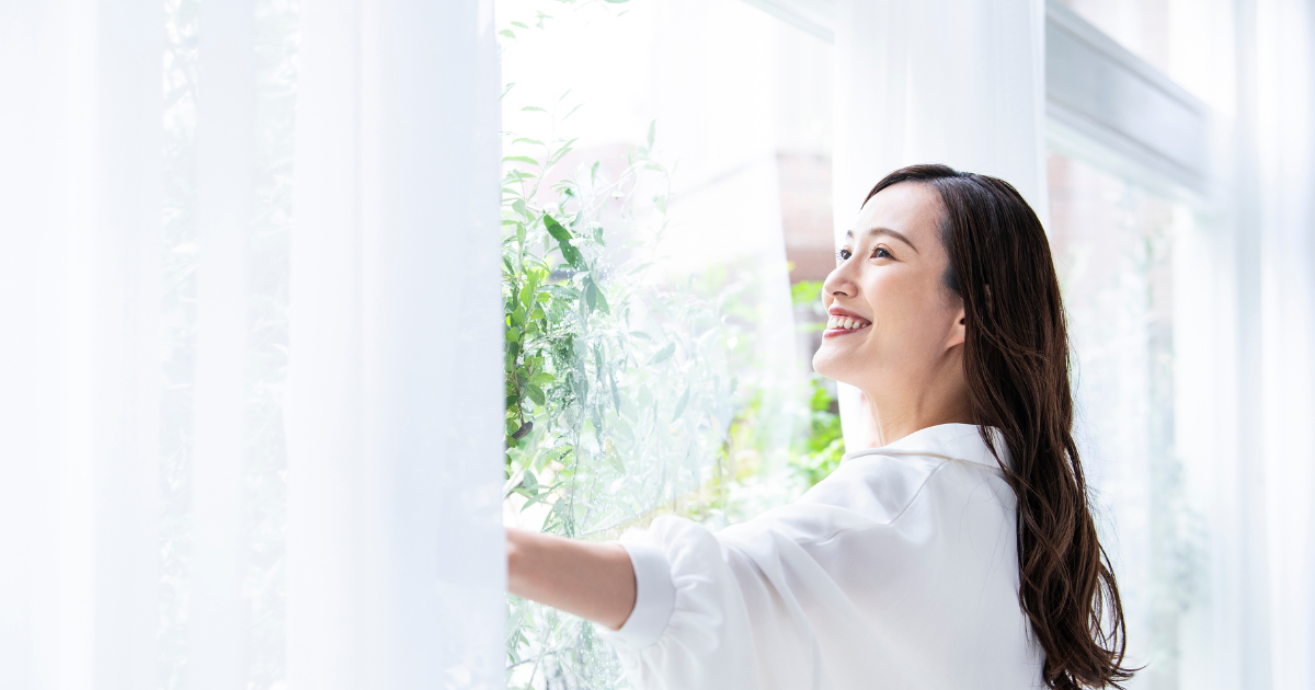 窓辺でカーテンを開けながら外を見て笑う女性