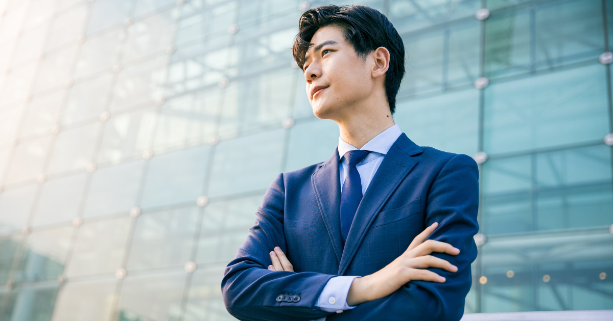 ガラス張りのオフィスビルを背景に、腕を組んで立つスーツ姿のビジネスパーソン
