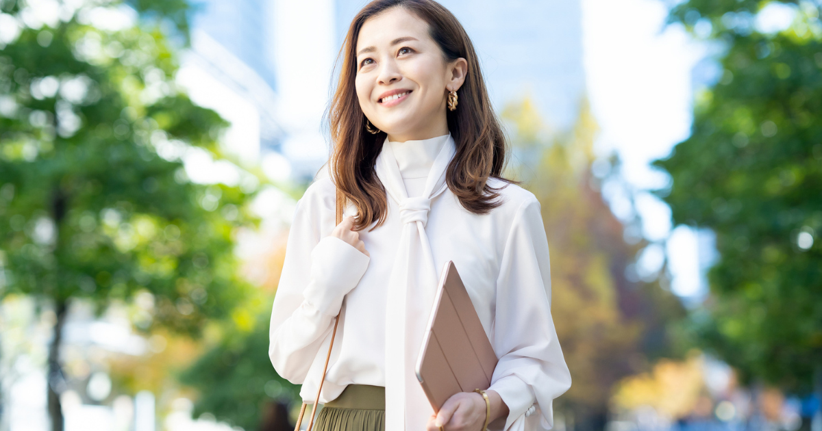 背景に木々があり、書類を抱えて微笑む女性