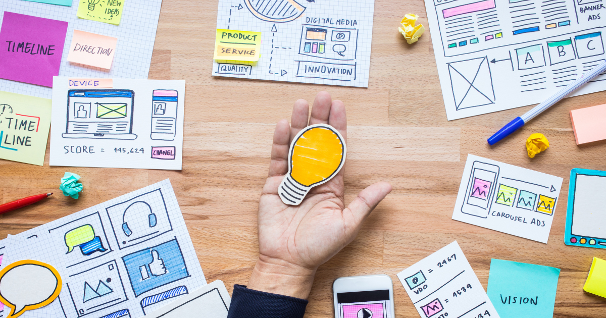 机の上に付箋や図のメモが並び、中央の手に小さな電球のイラストが置いてある