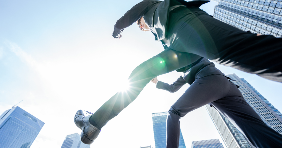 ビルの合間で、前方に向かって力強く踏み出すスーツ姿の人物の足元