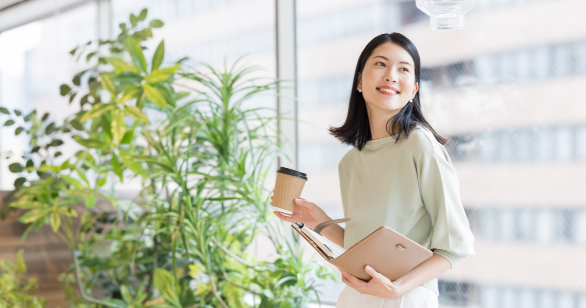 観葉植物のそばで、手帳とコーヒーを持って立つ笑顔の女性