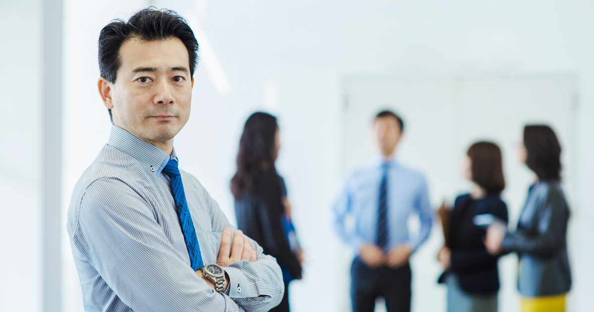 社内チームを背後に、腕を組み、カメラを見て立つビジネスパーソン