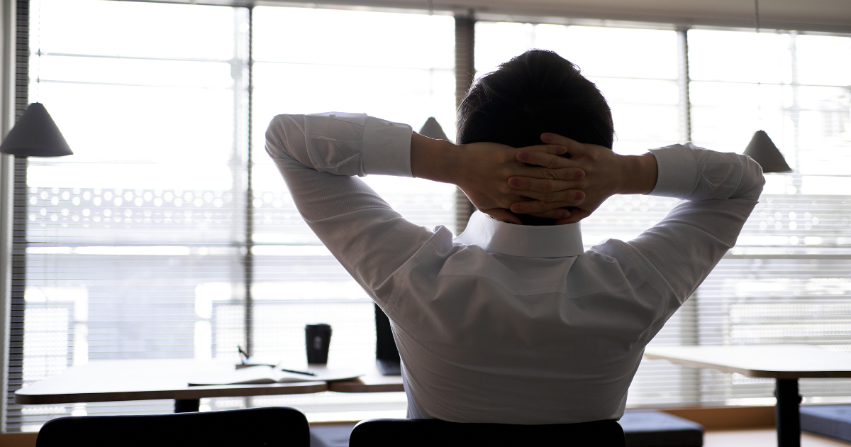 窓の前で椅子に座り、頭の後ろで手を組む男性の後ろ姿