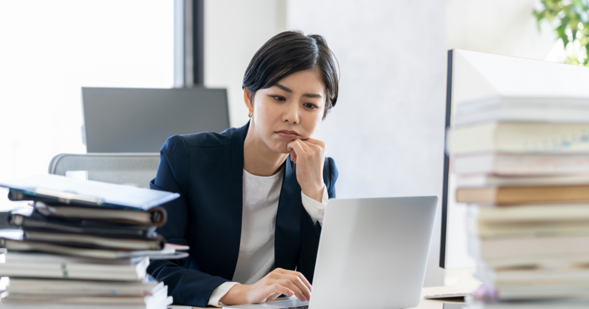 資料が積まれたデスクで、ノートパソコンを見ながら考え込む女性