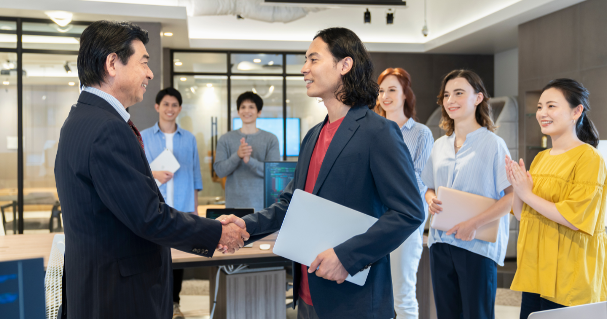 同僚たちが見守る中で握手をする二人の男性