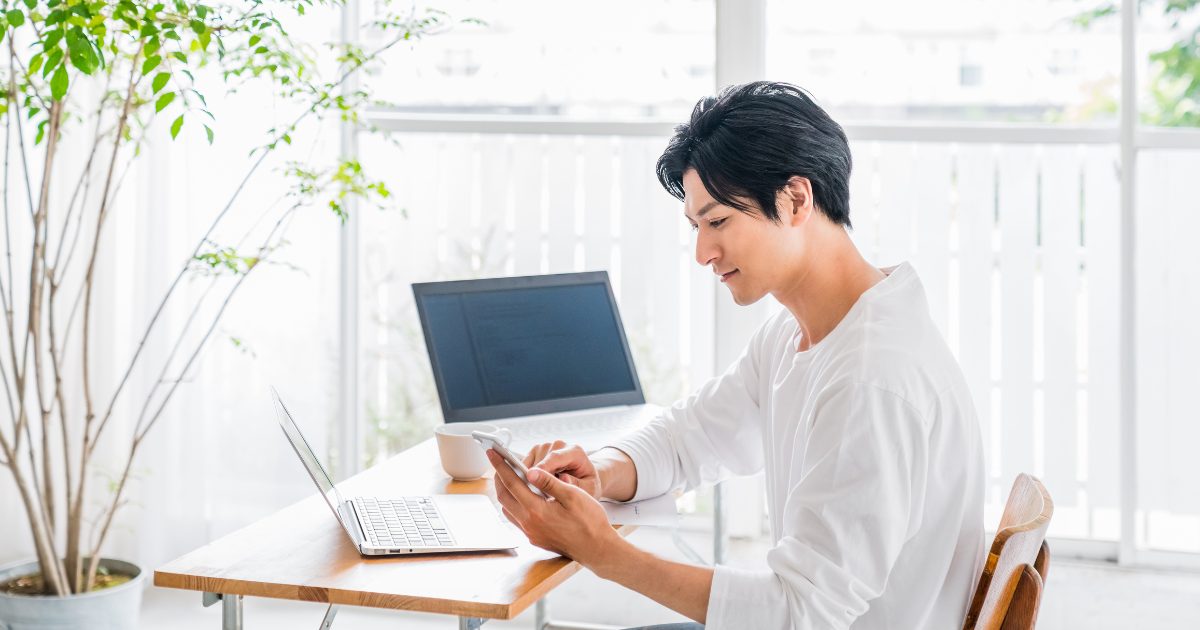 デスクでノートパソコンを開き、スマートフォンを確認する男性