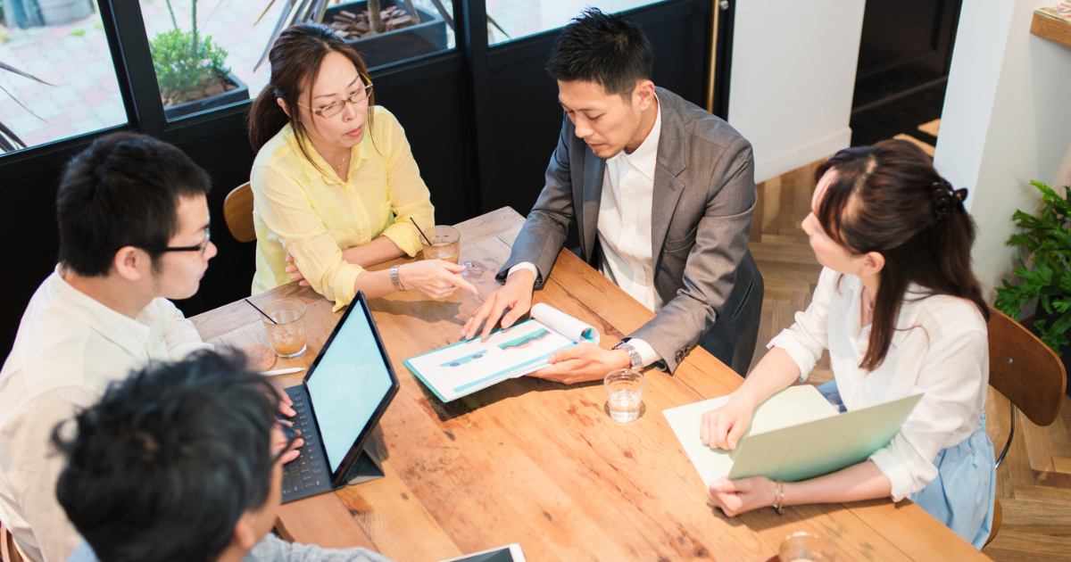 カフェ風のスペースで資料を囲みながら話し合う男女4人のグループ