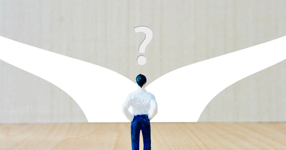 ビジネスマンの人形が2つに分かれた道の前に立っていて、クエスチョンマークが浮かんでいる