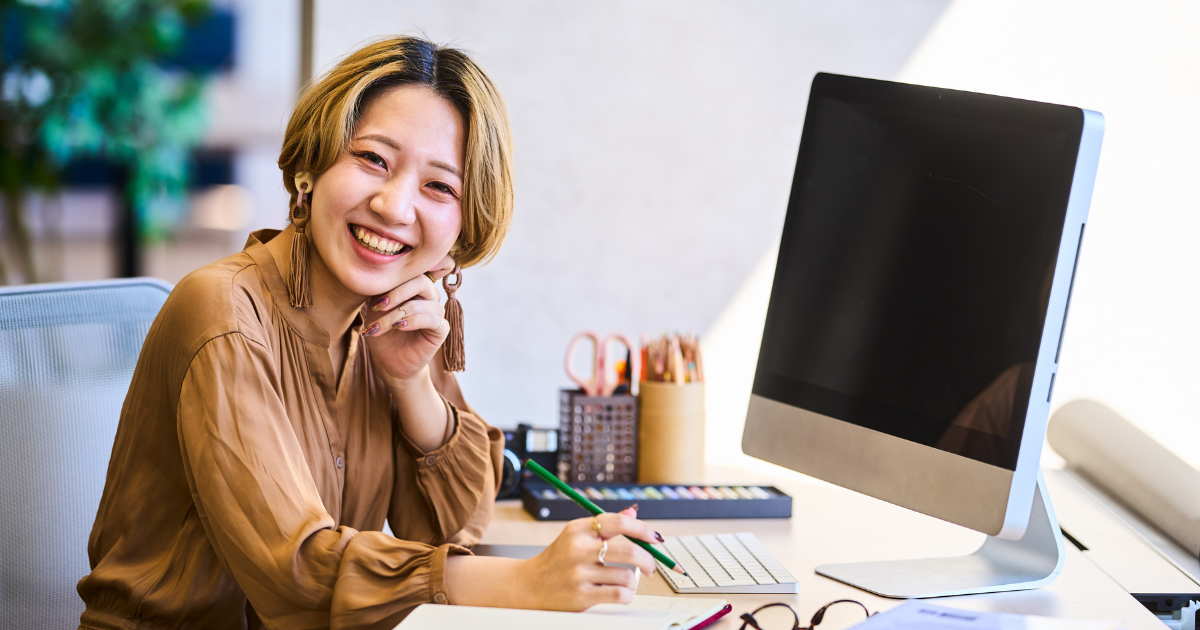 デスクトップパソコンの前で頬杖をつき、笑顔を見せる女性