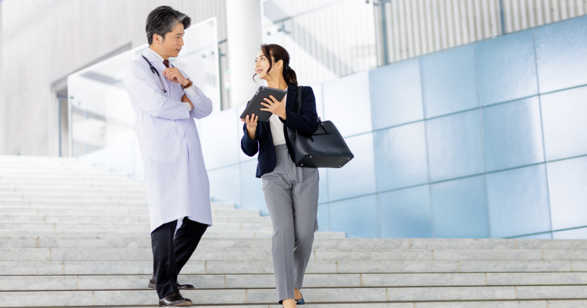 屋外の階段を歩きながら話す白衣の男性とスーツ姿の女性