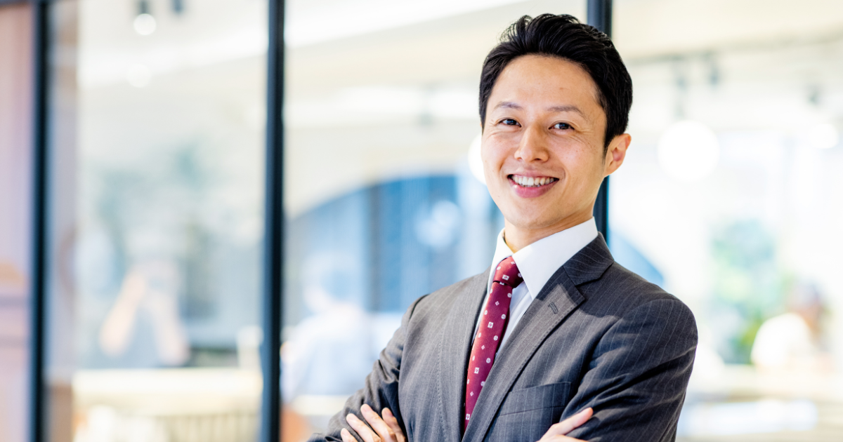 腕組みをして笑顔で立つスーツ姿の男性