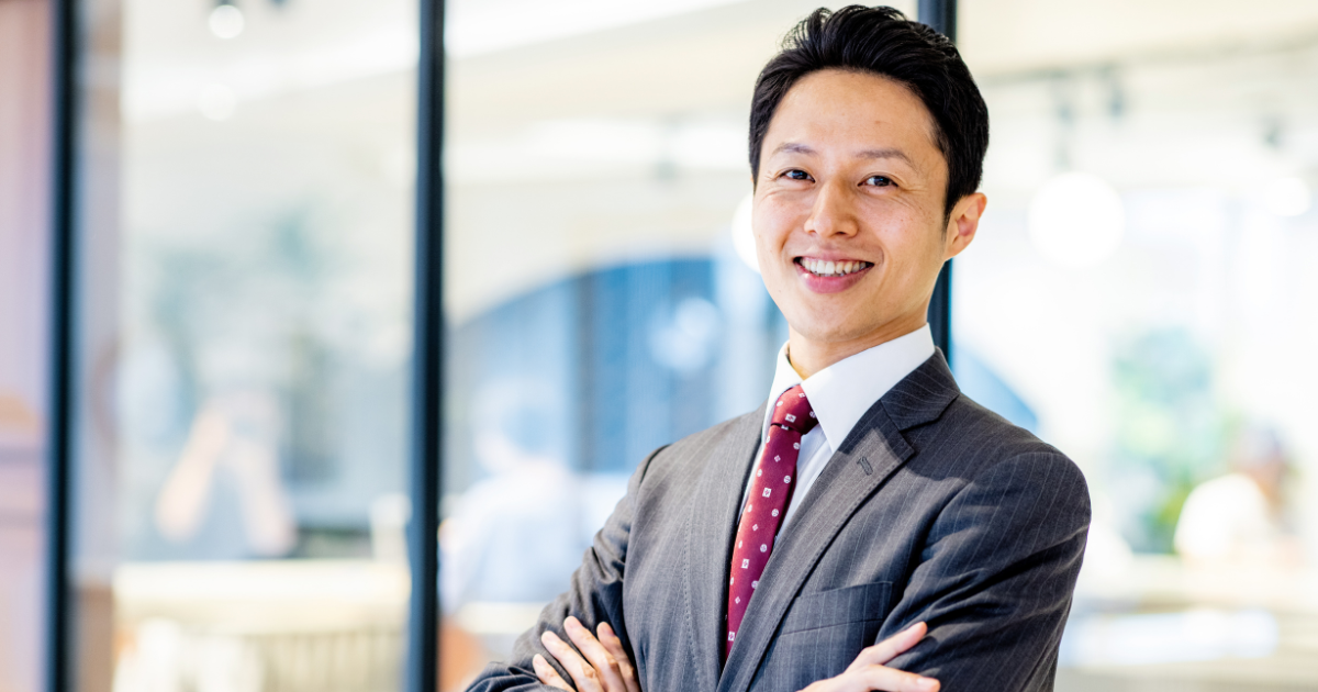 明るいオフィスで腕を組み、笑顔で立つスーツ姿の男性
