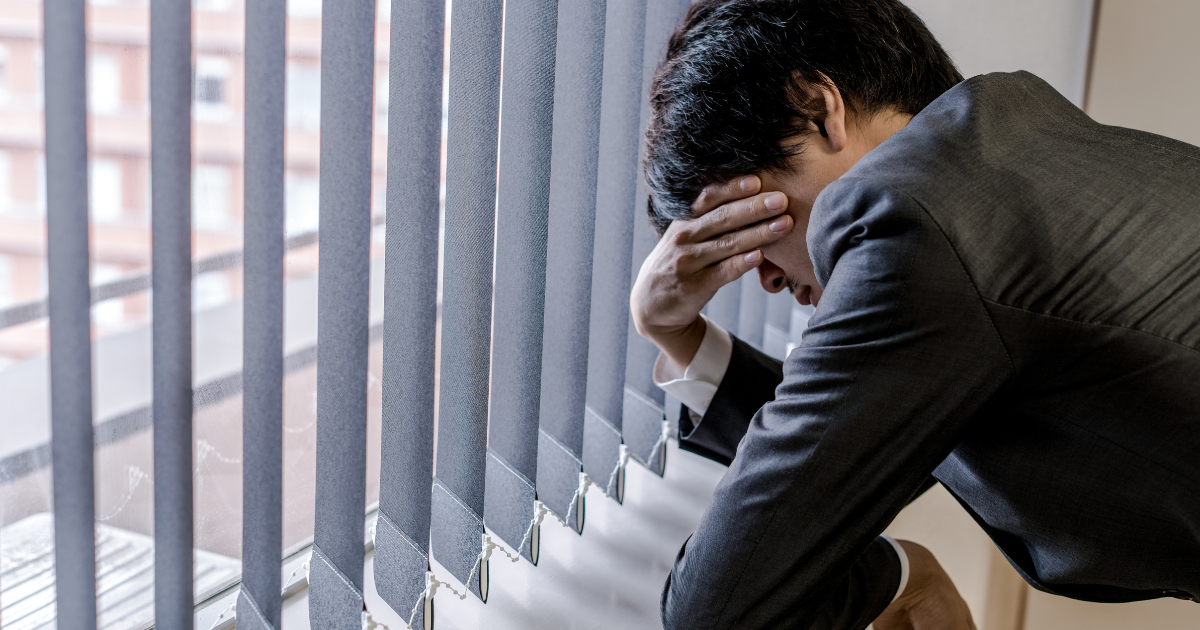 ブラインドの横で頭を抱えて悩むスーツ姿の男性
