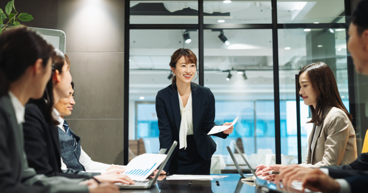 会議室で立ち上がって笑顔で進行する女性と、着席して話を聞く参加者たち
