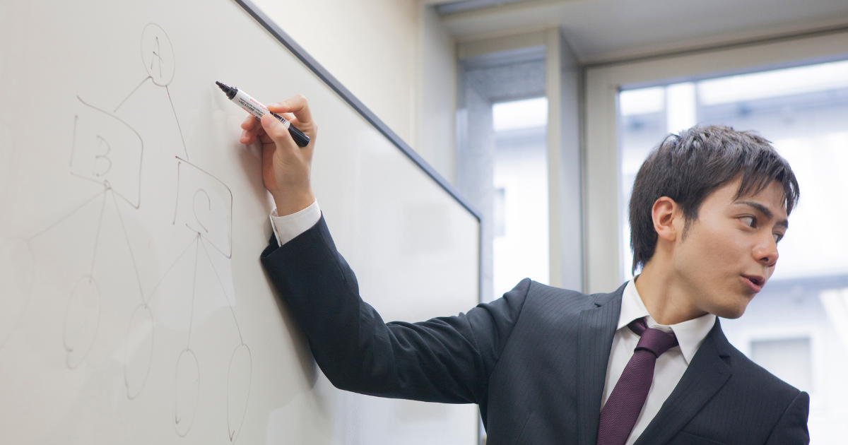 ホワイトボードにマーカーで書き込みながら振り返るスーツ姿の男性